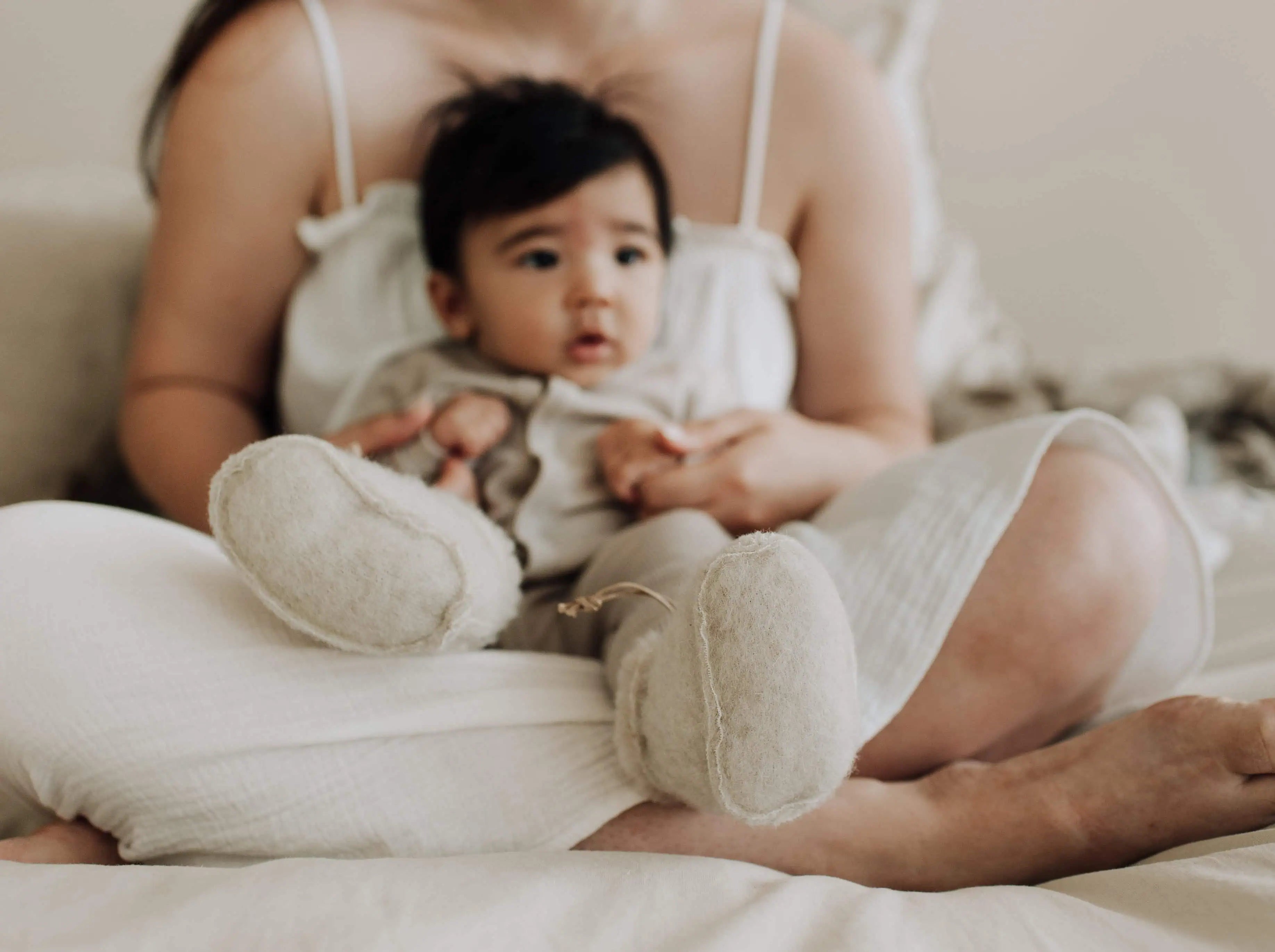 Baby met zwart haar en wollen sloffen die bij zijn moeder op schoot zit. De moeder zit in een kleermakerszit