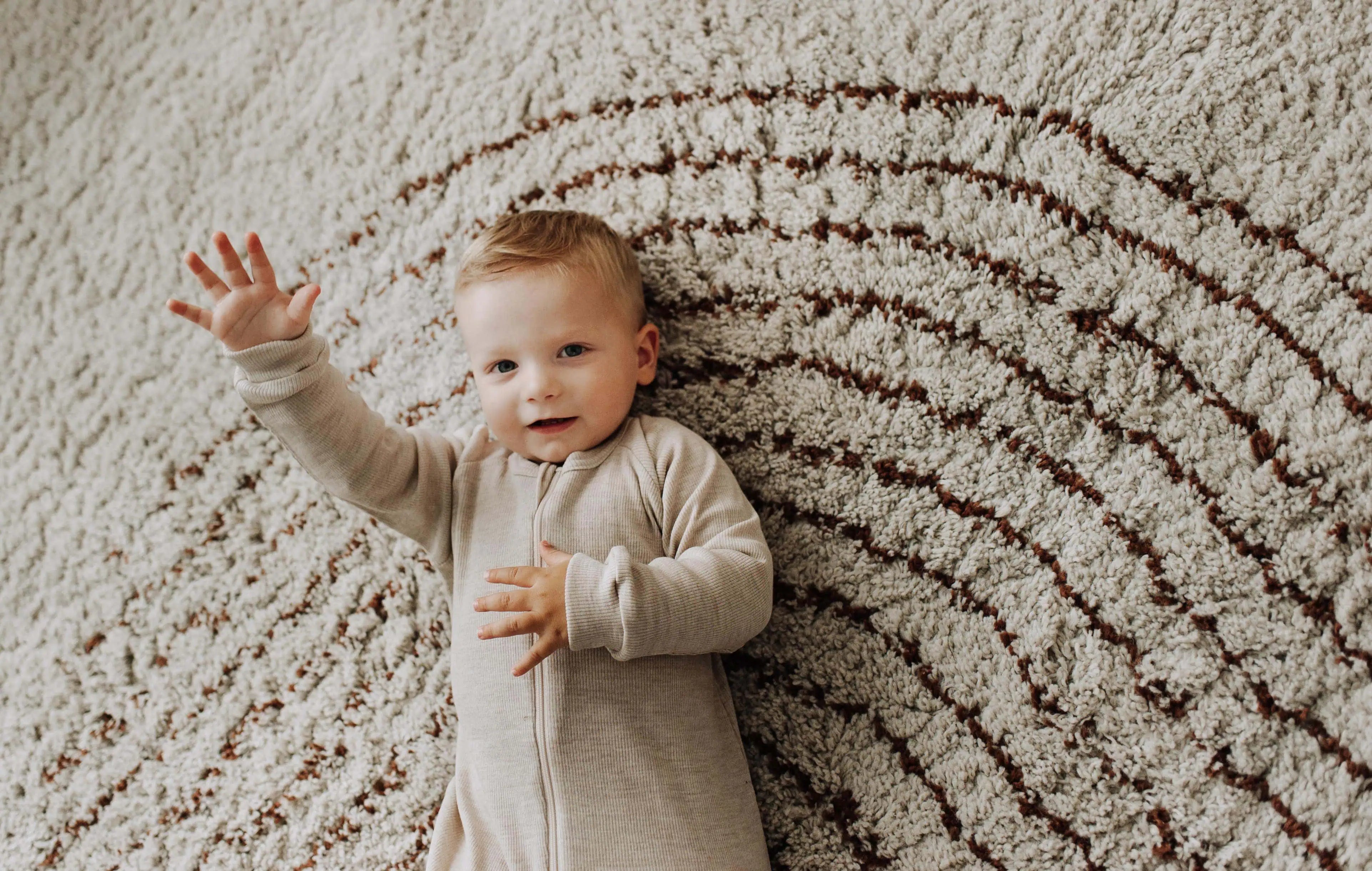 Baby in wollen slaapzak die op een tapijt ligt en zijn hand opsteekt